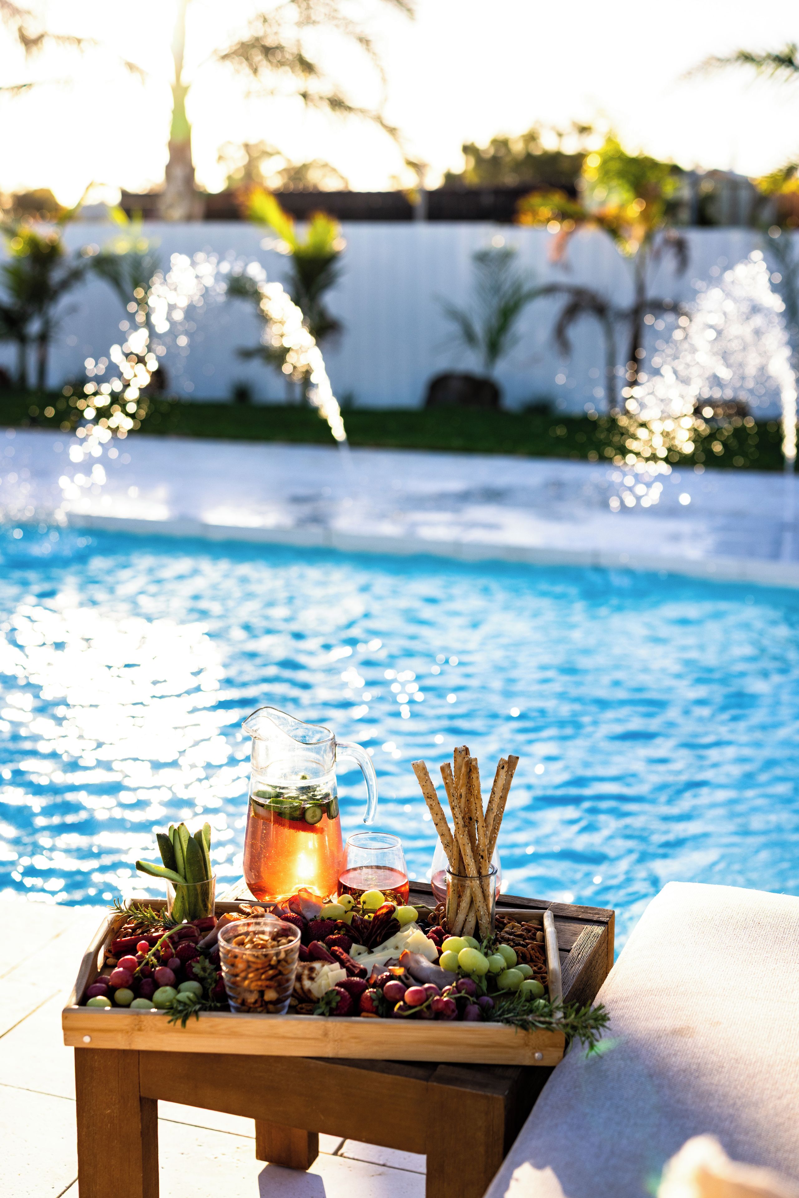 Ein Tisch am Pool mit einer Platte Obst und Käse, einer Karaffe und einem Glas Drink.