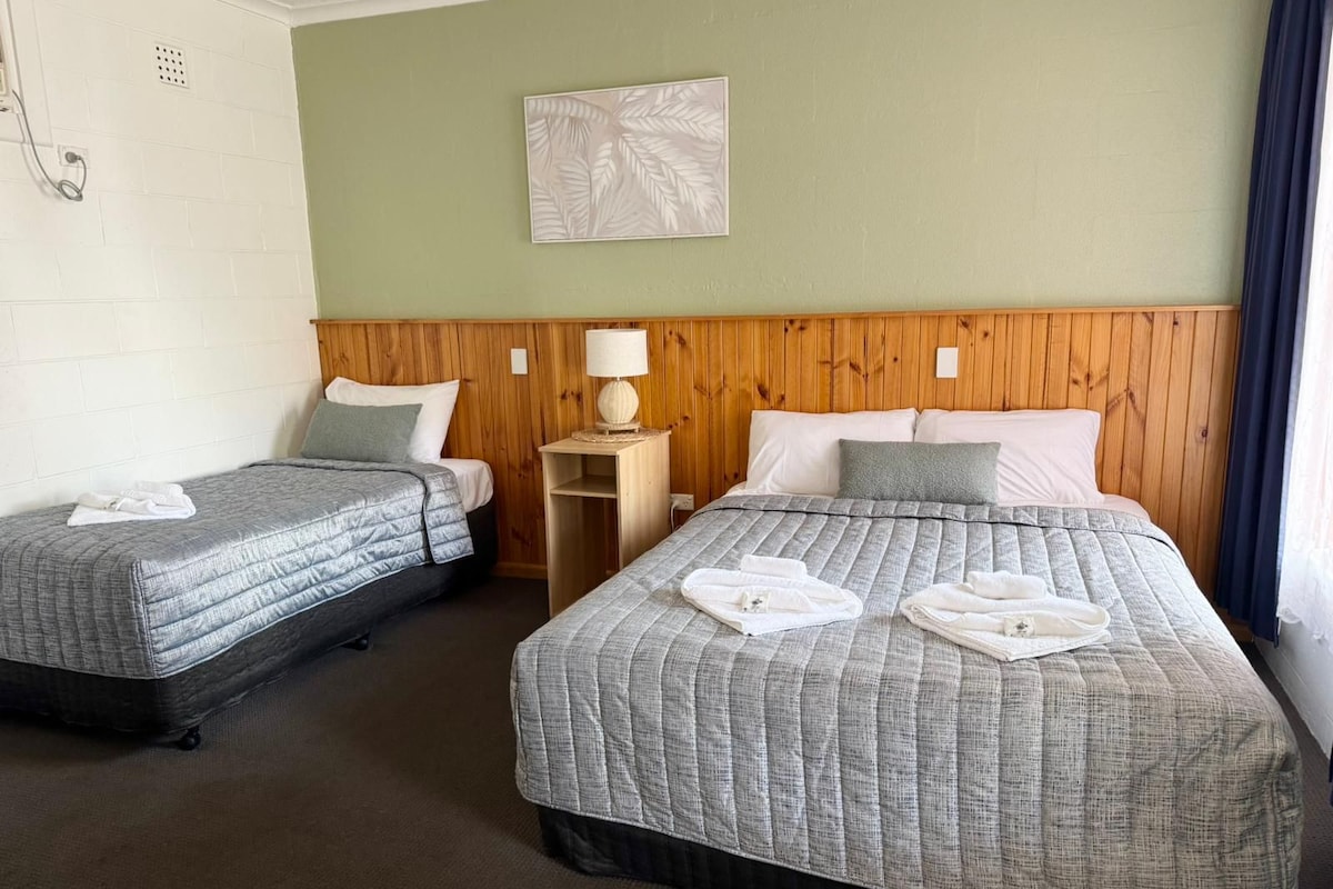 A hotel room with two beds, a wooden headboard, a small bedside table with a lamp, and a wall-mounted artwork.