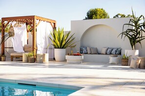 A poolside area with a wooden gazebo, seating, potted plants, and a white wall with an arched window.