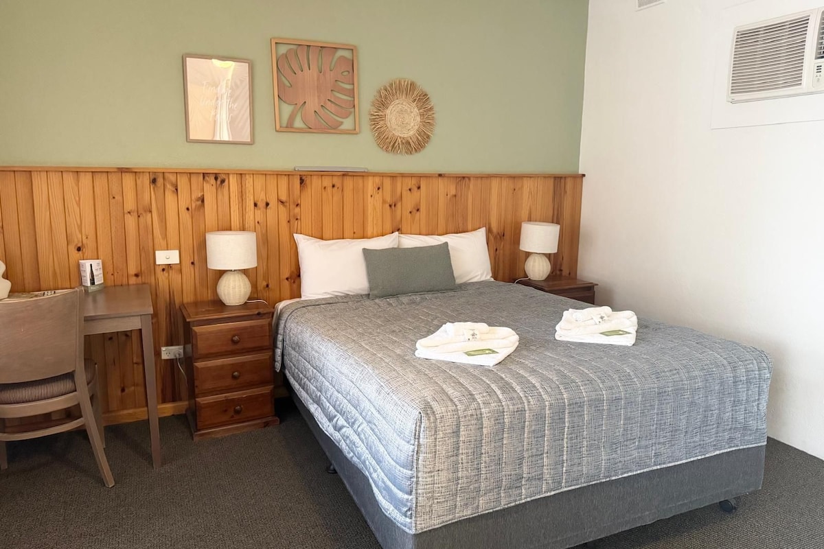 A hotel room with a wooden headboard, a bed with a grey quilt, two bedside lamps, a small desk, and a chair.