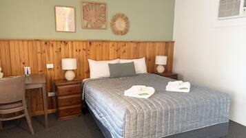 Una habitación de hotel con cabecera de madera, cama con colcha gris, dos lámparas de noche, un escritorio pequeño y una silla.