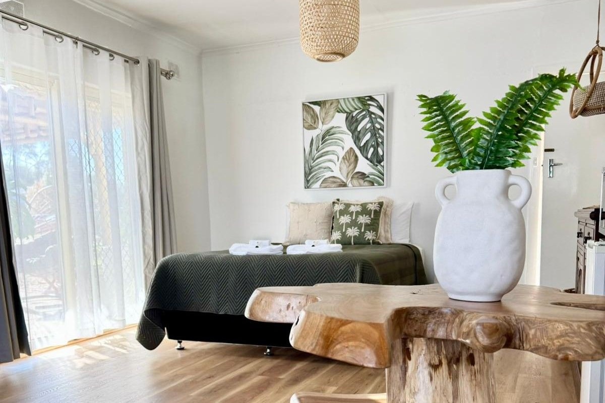 Un dormitorio con una cama, un ventanal con cortinas, una lámpara colgante, una obra de arte en la pared y una mesa de madera con una planta.