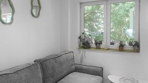 Um quarto com um sofá cinzento, uma pequena mesa branca e uma janela com plantas.