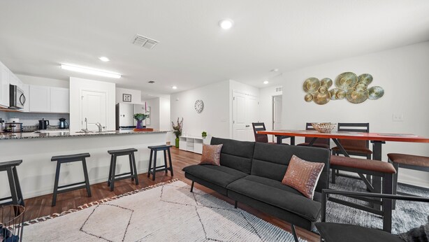 Una cocina moderna con armarios blancos, una isla central con taburetes, una zona de comedor con mesa y sillas de madera, y una sala de estar con un sofá oscuro y alfombra estampada.