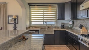 A modern kitchen with dark cabinetry, a granite countertop, a built-in oven, and a window with blinds. - Tortuga TBB 2401 (San José del Cabo)