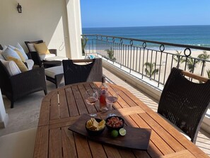 Una mesa de madera con una botella de vino, dos copas, un bol de patatas fritas y una lima en un balcón con vistas a la playa.
