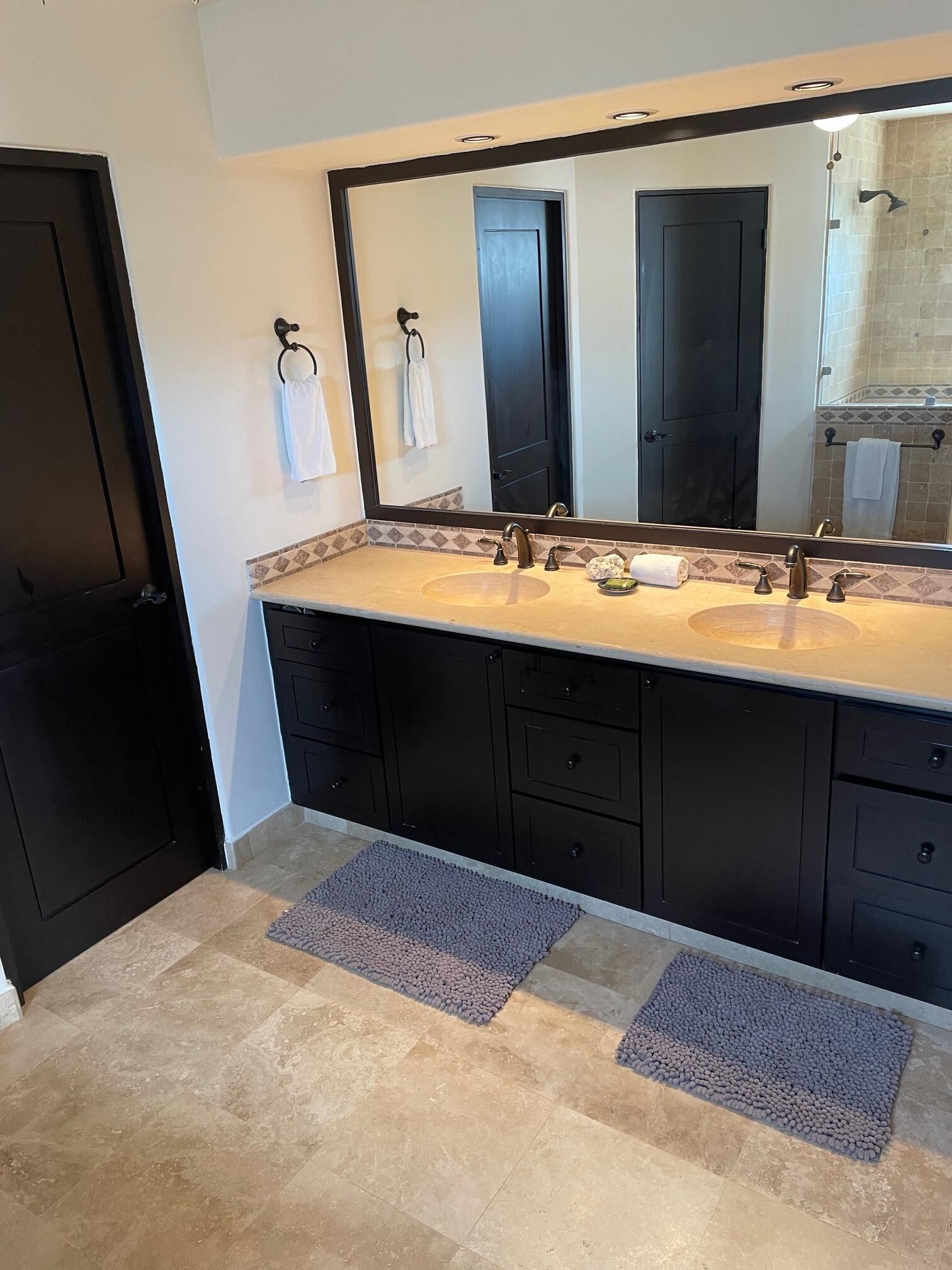 A bathroom with a large mirror, two sinks, black cabinets, and two small rugs on the floor.