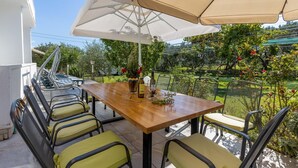 An outdoor patio with a wooden table and chairs, a large umbrella, and a view of a garden with trees and flowers. - Beautiful apartment in Banjol with WiFi (Banjol)