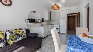 Un espacio habitable compacto con sofá, una mesa de comedor con fruta, una cocina con electrodomésticos blancos y un lámpara colgante.