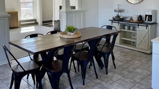 A dining area with a wooden table and six chairs, a hanging light fixture, a kitchen counter with a coffee maker, and a large mirror on the wall.