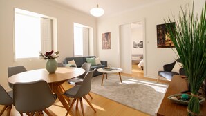 A modern living room with a dining area, featuring a round wooden table, grey chairs, a grey sofa, and a large green plant. - O Beco - Apart In The Heart Of The City (Viana do Castelo)