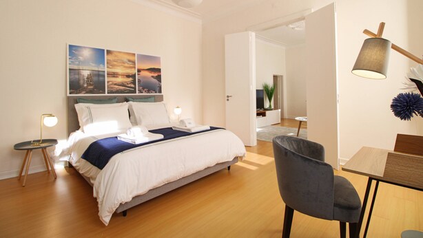 A modern hotel room with a neatly made bed, a dining area with a table and chairs, and a hallway leading to another room. - O Beco - Apart In The Heart Of The City (Viana do Castelo)