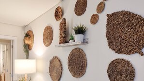 Habitación con una pared blanca adornada con varios tapices. Se observa una pequeña estantería con una planta y una maceta blanca. En la esquina, una lámpara de pie y otra planta en maceta son visibles.