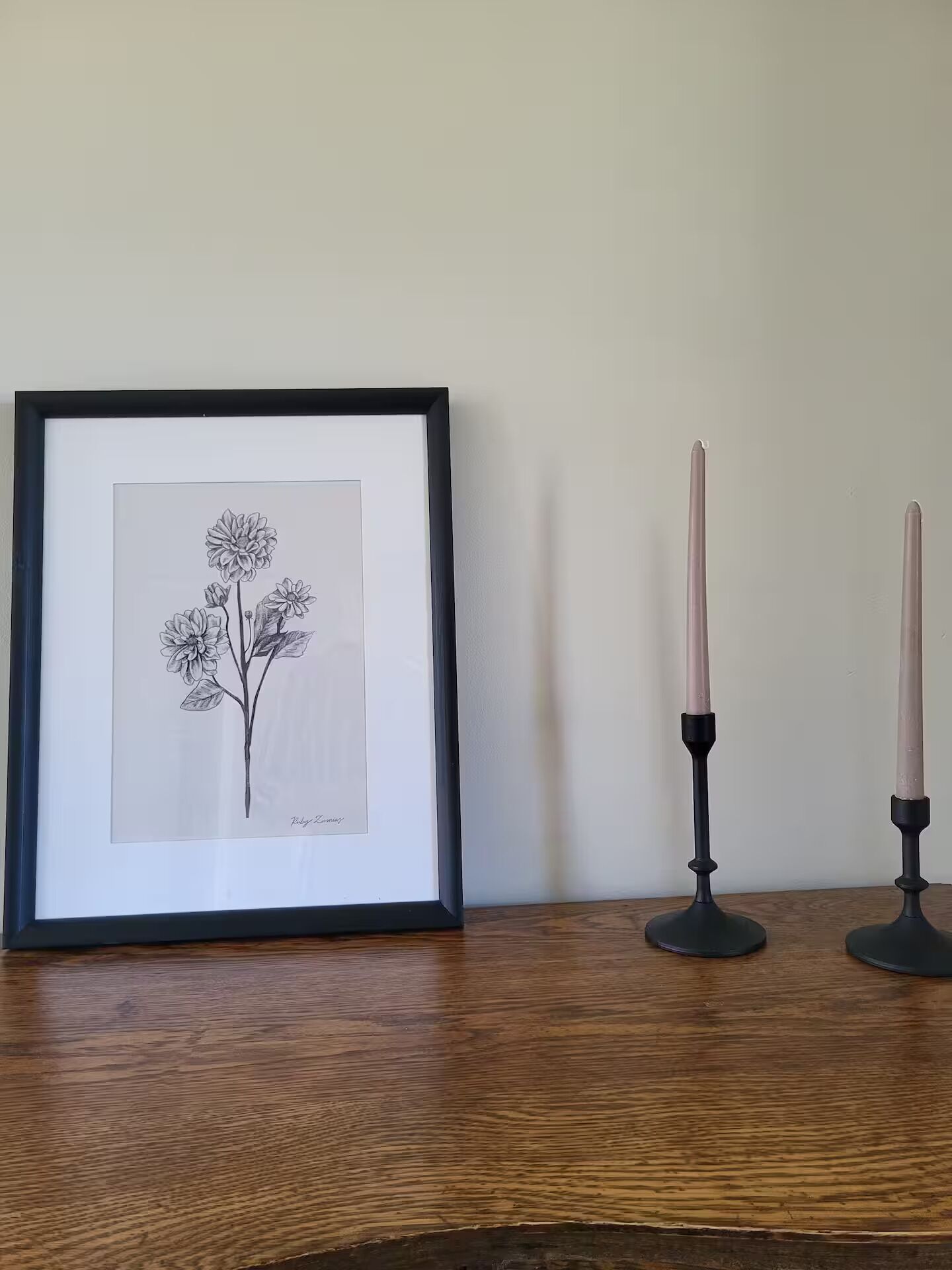 Een ingelijste botanische tekening op een houten oppervlak, met aan weerszijden twee zwarte kandelaars.