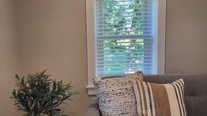 Un salón con un sofá gris, un florero de vidrio con plantas verdes y una ventana con persianas.