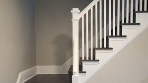 Una escalera con escalones oscuros y barandillas blancas, que conduce a un rellano con una cesta pequeña blanca y una rejilla de ventilación.