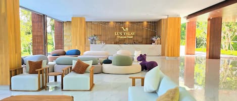 A modern hotel lobby with a reception desk, plush seating, and a view of greenery outside.