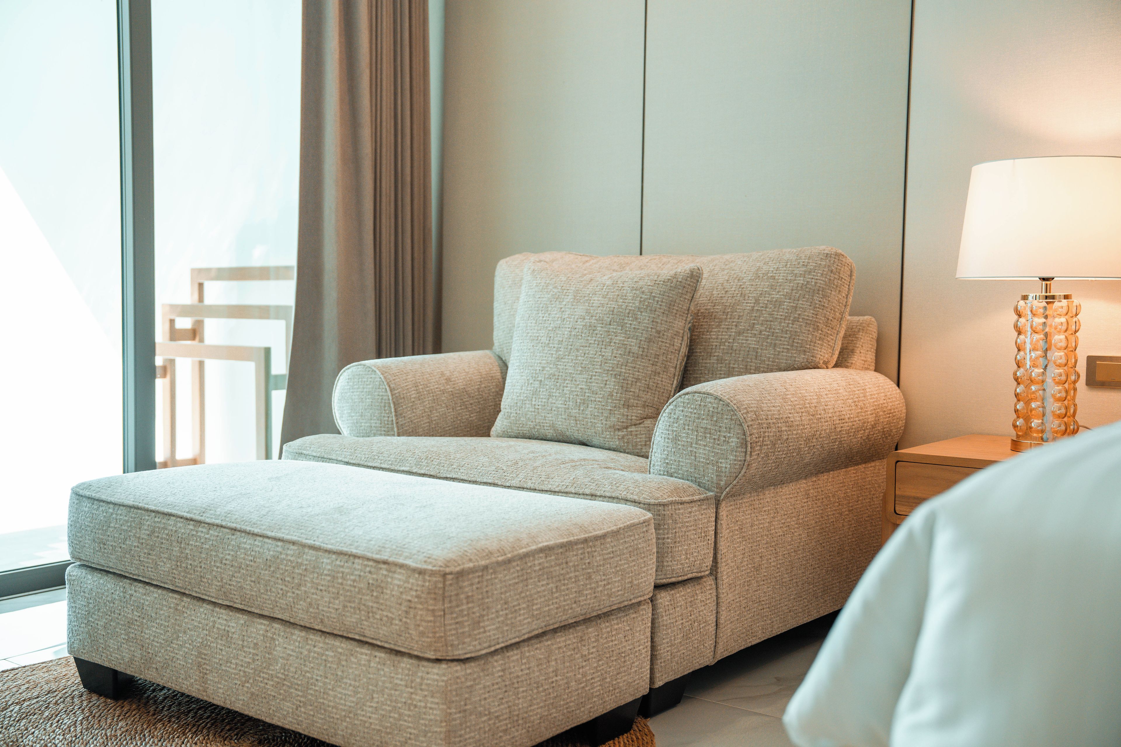 Ein modernes Hotelzimmer mit einem beigen Sofa, einem Nachttisch mit Lampe und einem großen Fenster mit Vorhängen.