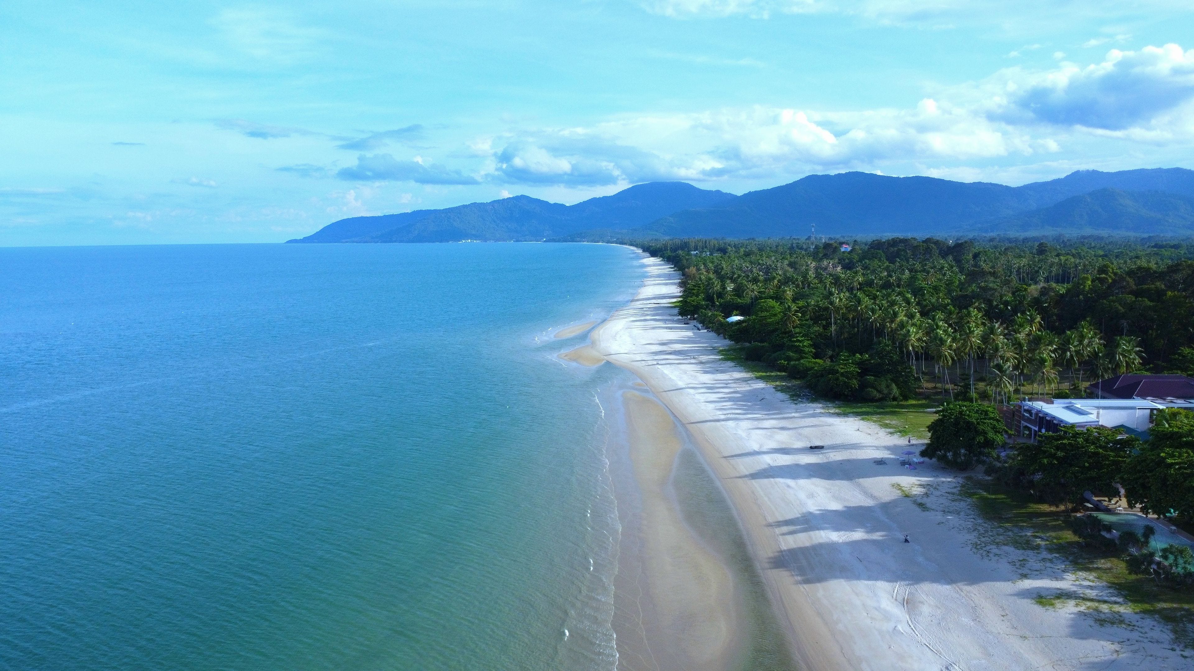 An aerial view of a beach with a long stretch of sandy shore, palm trees, and mountains in the background.