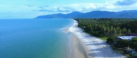 An aerial view of a beach with a long stretch of sandy shore, palm trees, and mountains in the background.