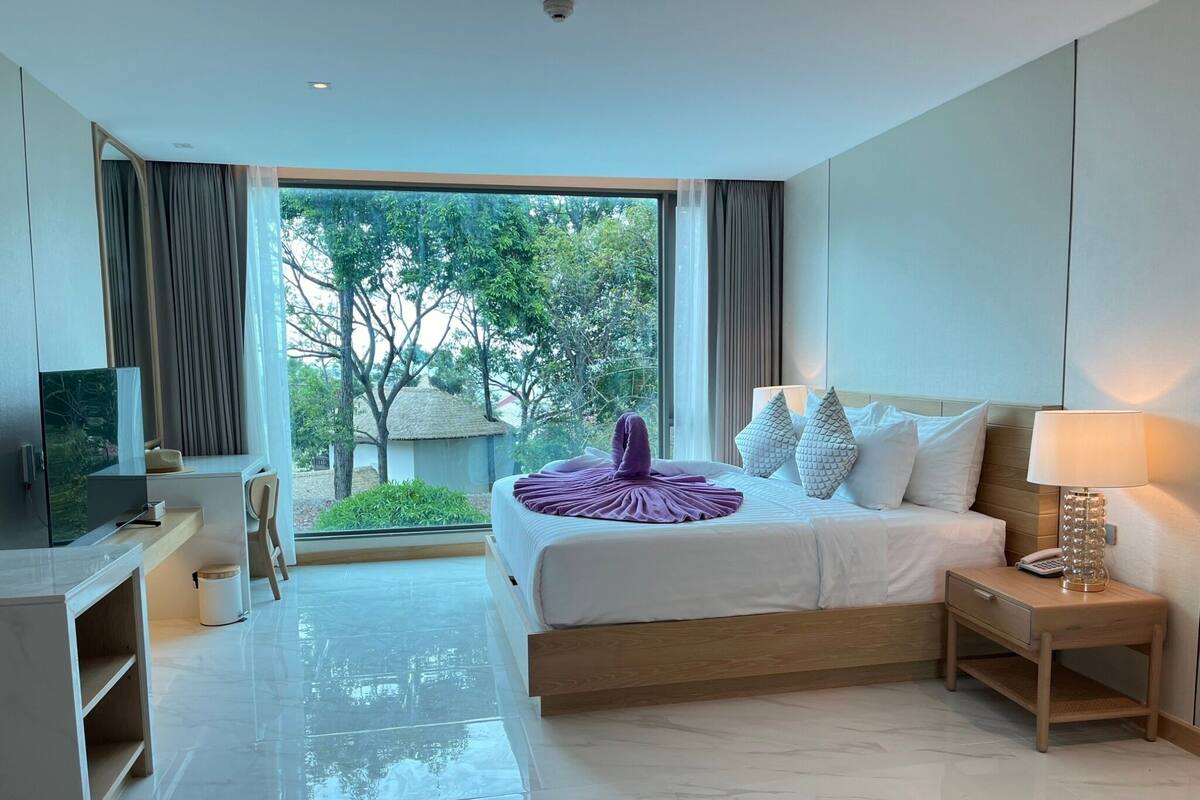 Une chambre d’hôtel moderne dotée d’un grand lit, d’un bureau, d’une télévision et offrant une vue sur la verdure environnante.