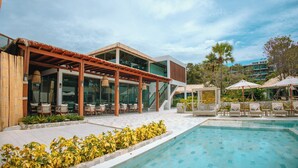 Zona junto a la piscina con un área de estar cubierta, mobiliario exterior y vista despejada al estanque.