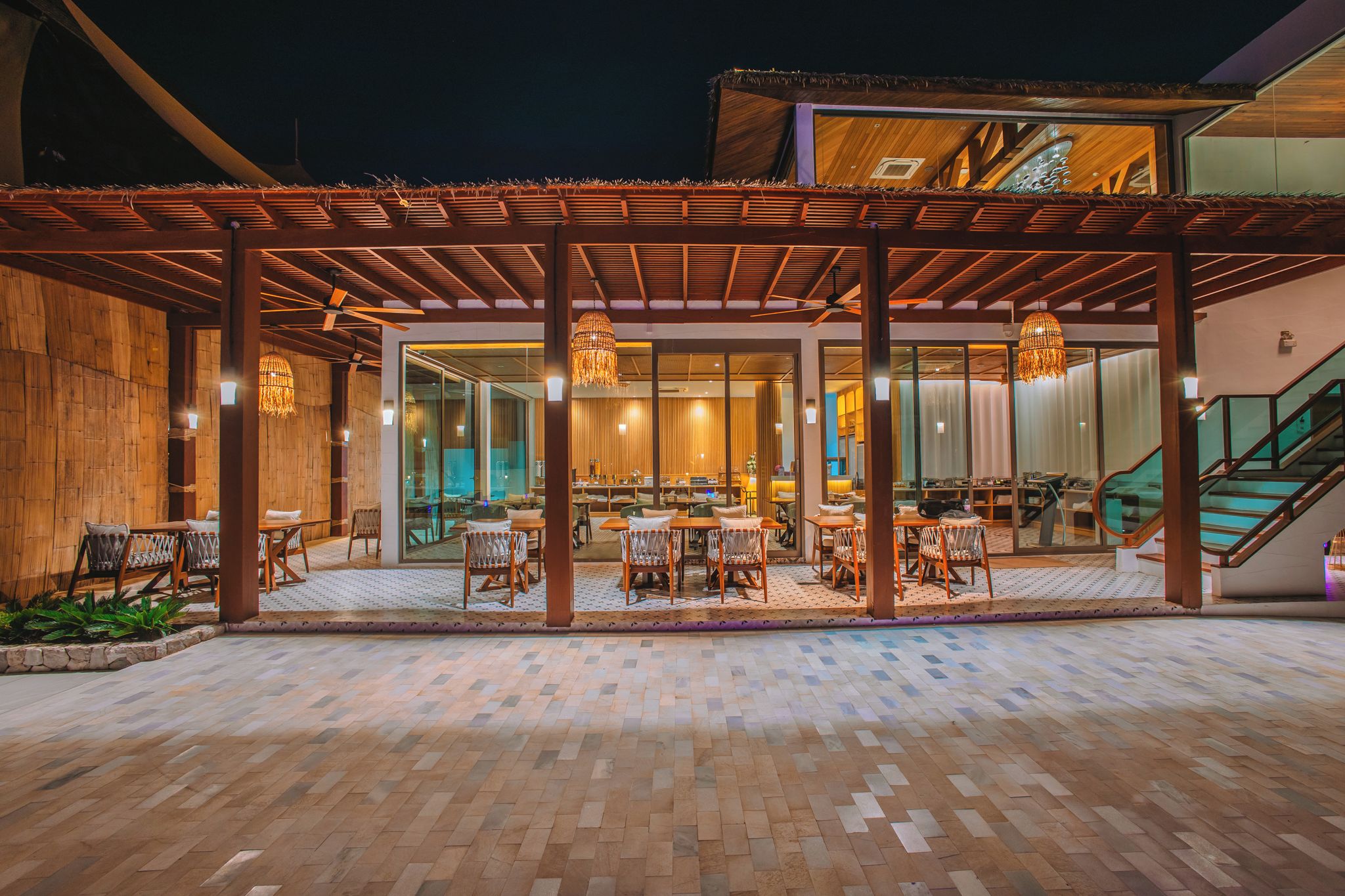 An outdoor dining area with wooden tables and chairs, a covered seating area, and a staircase leading to an upper level.