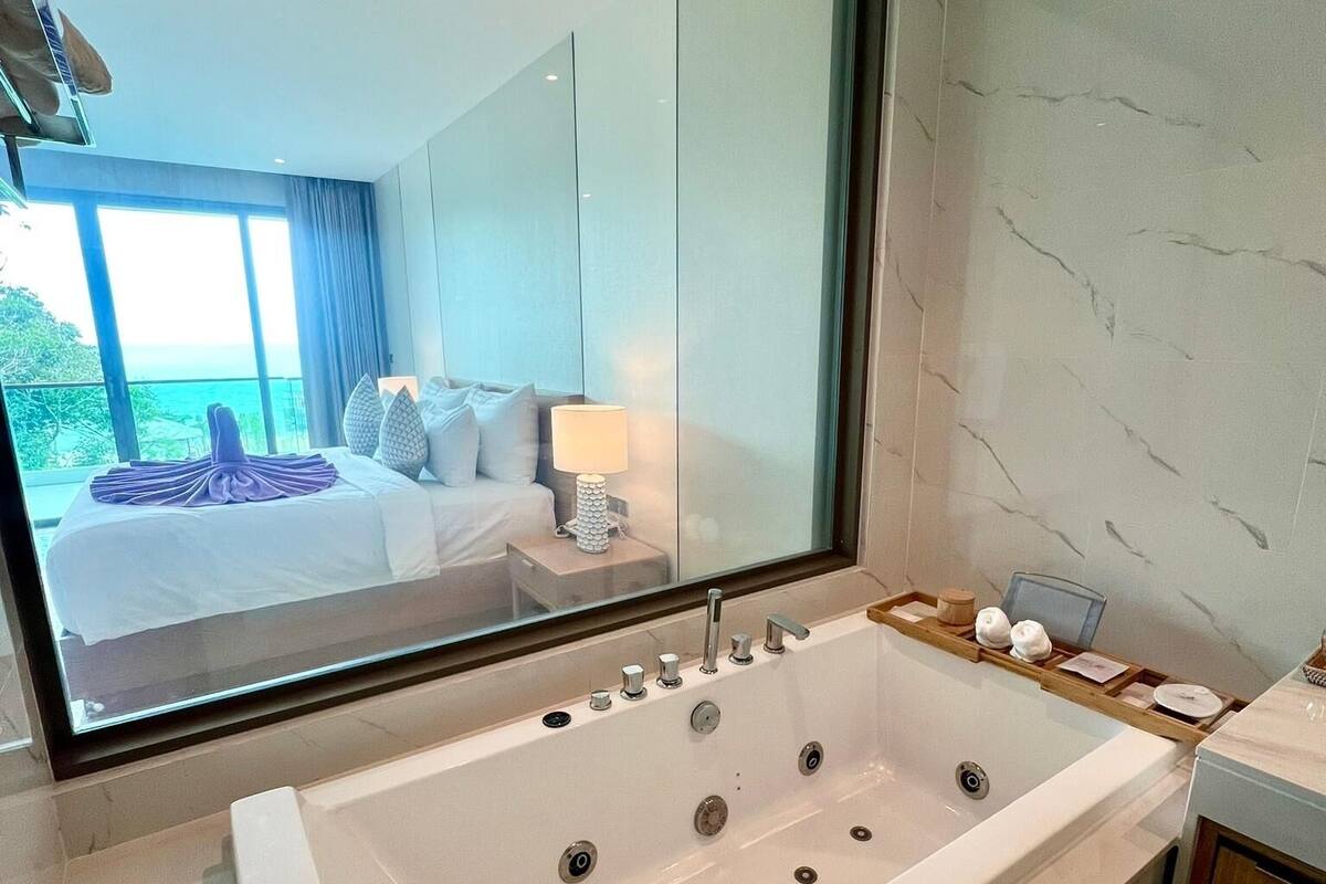 Une salle de bain moderne avec une grande baignoire, un miroir et une vue sur une chambre à coucher avec un lit et une fenêtre.