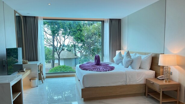 A modern hotel room with a large bed, a desk, a TV, and a view of greenery outside. - Luxtalay Khanom Hotel (Khanom)