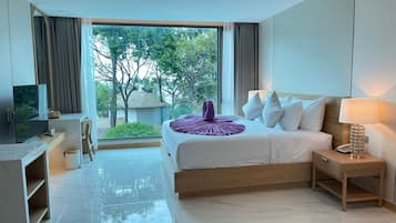 A modern hotel room with a large bed, a desk, a TV, and a view of greenery outside.