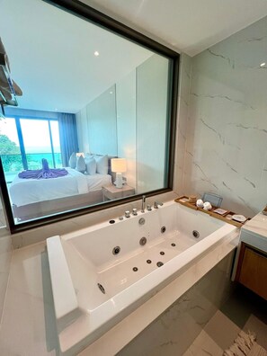 A modern bathroom with a large bathtub, a mirror, and a view of a bedroom with a bed and a window. - Luxtalay Khanom Hotel (Khanom)