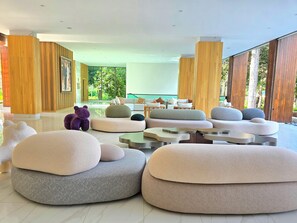 A modern lounge area with multiple seating options, including large, round ottomans and a sofa, arranged in a spacious, well-lit room with large windows offering a view of greenery outside.