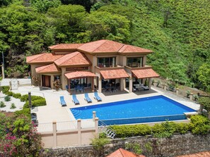 A large house with a swimming pool, multiple outdoor seating areas, and a lush green hillside in the background.