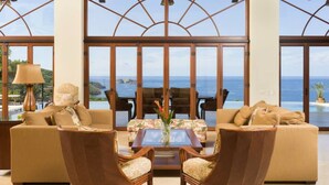 A spacious living room with a large window offering a view of the ocean. There are two sofas, a coffee table, and two armchairs.