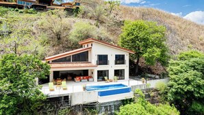 Ein modernes, zweistöckiges Haus mit Swimmingpool, umgeben von üppiger Vegetation und Hügeln.