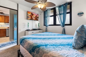 A bedroom with a bed, a ceiling fan, a painting, a window with curtains, and a kitchen visible through an open door. - Kona Bali Kai#307 (Kailua-Kona)