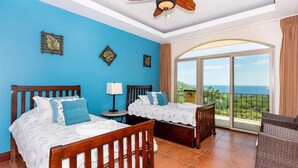 A hotel room with two wooden beds, a ceiling fan, a nightstand, and a view of the ocean. - Playa Hermosa Hillside Villa • 5BR • Sleeps 12 • Infinity Pool & Bay View • Casa Ilan Ilan (Playa Hermosa)