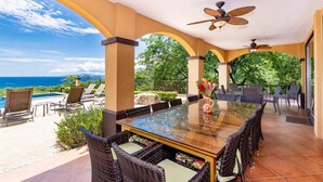 A covered outdoor dining area with a glass table, wicker chairs, and a view of the ocean. - Playa Hermosa Hillside Villa • 5BR • Sleeps 12 • Infinity Pool & Bay View • Casa Ilan Ilan (Playa Hermosa)