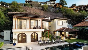 A two-story house with a swimming pool, a patio with lounge chairs, and a view of the surrounding hillside.