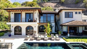 A two-story house with a swimming pool, outdoor seating, and a view of a hillside.