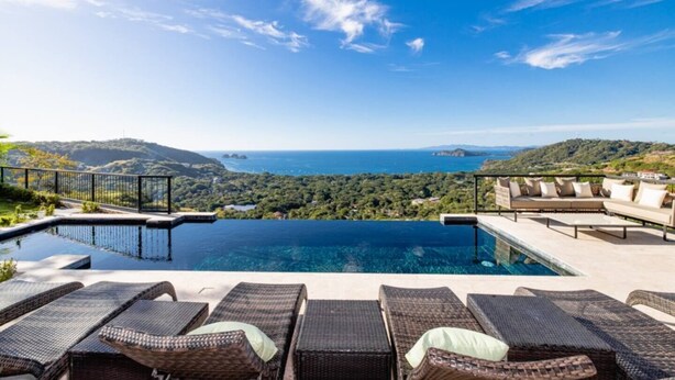 A infinity pool with a view of the sea and hills. There are lounge chairs and a sofa with cushions.