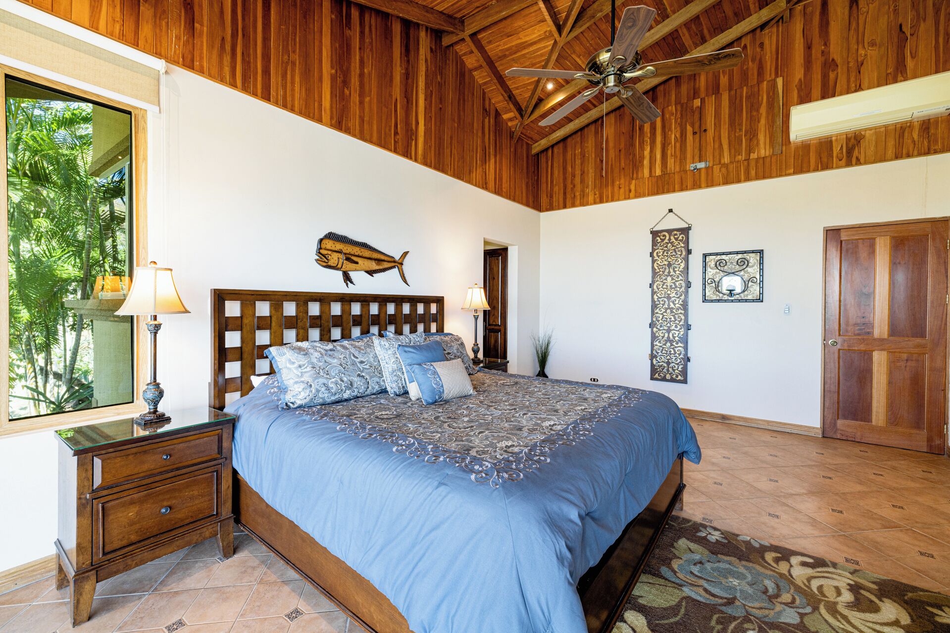 Une chambre avec un plafond en bois, un lit avec une literie bleue, une table de chevet avec une lampe, une décoration murale représentant des poissons et une porte.