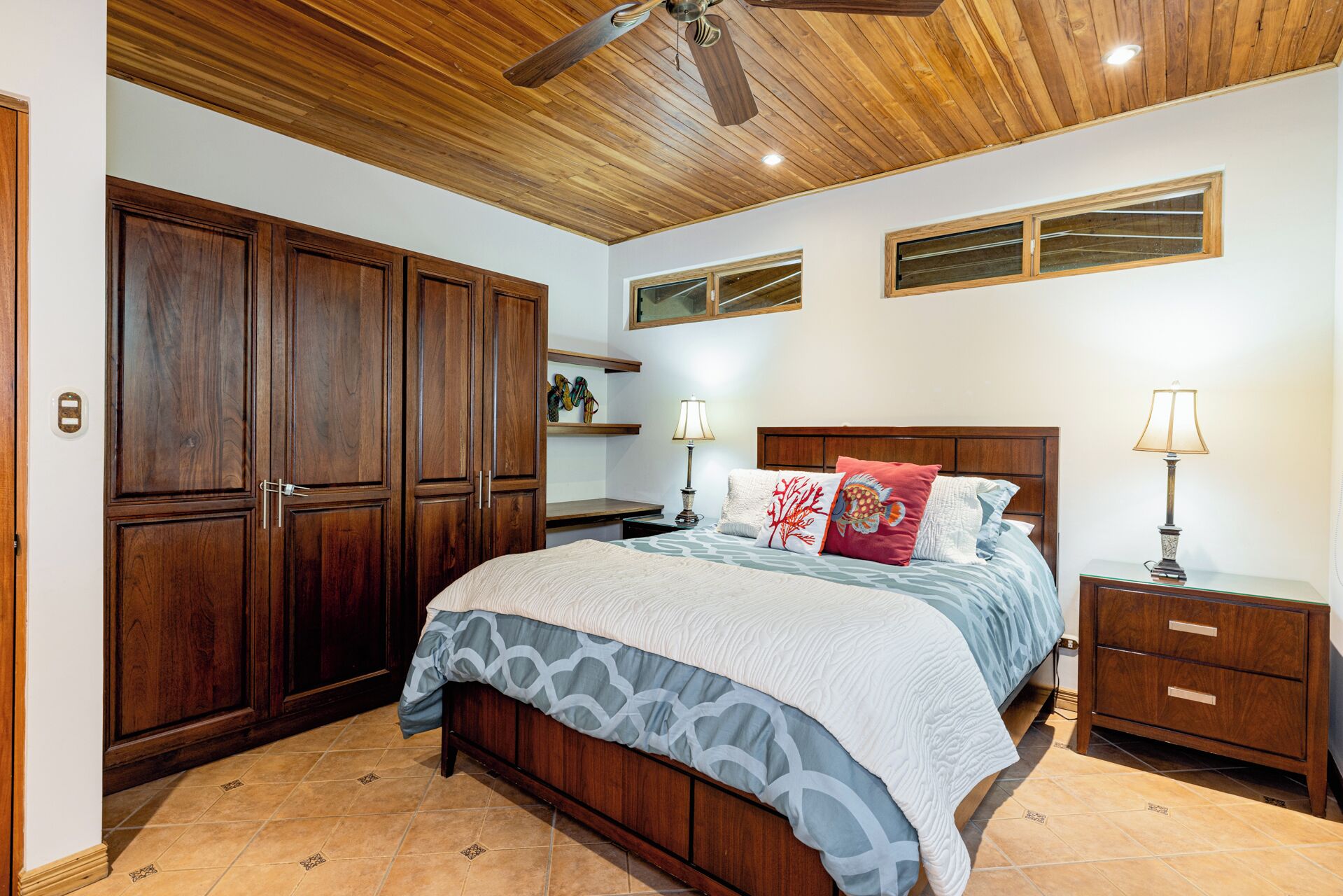 A bedroom with a wooden ceiling, a large bed with patterned bedding, two bedside tables with lamps, and a wardrobe.