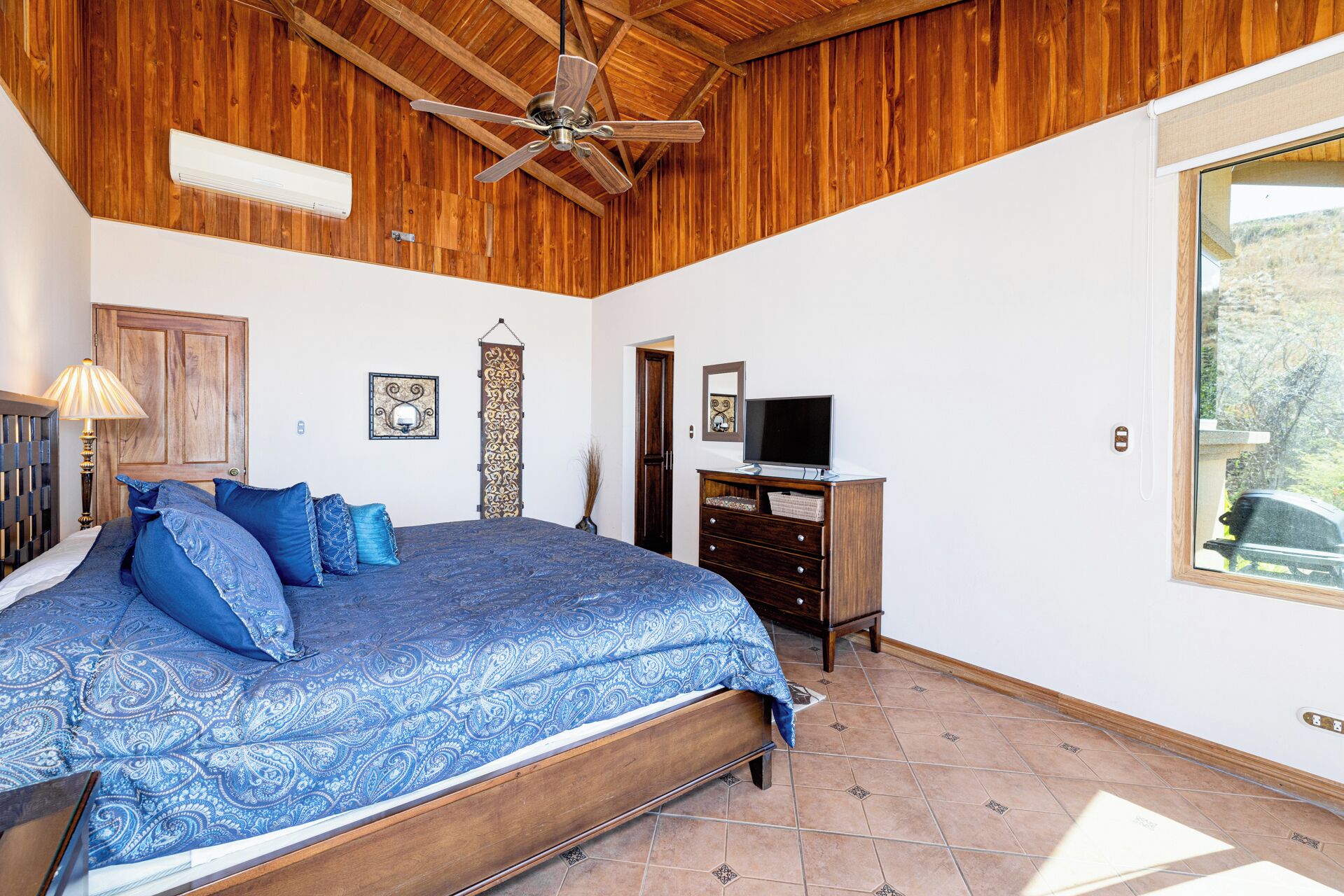 Un dormitorio con cama, techo de madera, ventilador de techo, televisor, cómoda y una ventana con vistas.