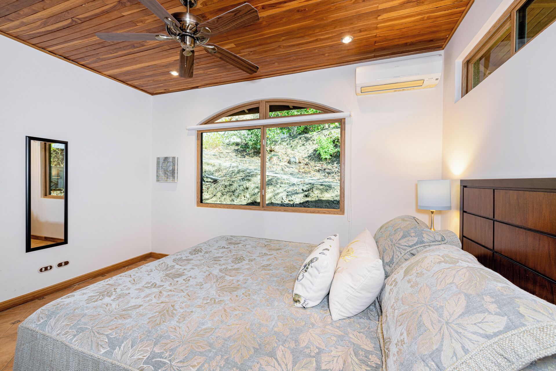 Un dormitorio con techo de madera, una cama grande con ropa de cama floral, una ventana con vista a follaje y un ventilador de techo.