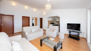 Un salon moderne avec un canapé, des fauteuils, une table basse en verre, un téléviseur et une cuisine en arrière-plan.