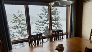 A dining area with a wooden table and chairs, large windows with a view of snow-covered trees. - 3-bedroom condo near mountain village on shuttle route (big sky)