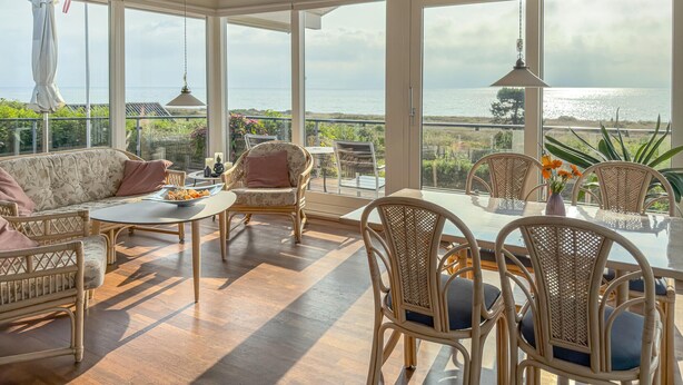A sunlit room with wicker furniture, large windows offering a view of the sea, and a table with a vase of flowers. - Stunning home in Ebeltoft with sauna (Ebeltoft)