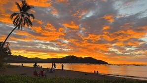 Eine Strandansicht mit Menschen, die am Sand entspannen, Booten auf dem Wasser und einem Sonnenuntergang mit orangefarbenen Wolken.
