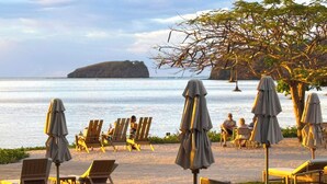 Eine Sitzgelegenheit am Strand mit Holzstühlen und Sonnenschirmen mit Blick auf ein ruhiges Meer und eine kleine Insel.